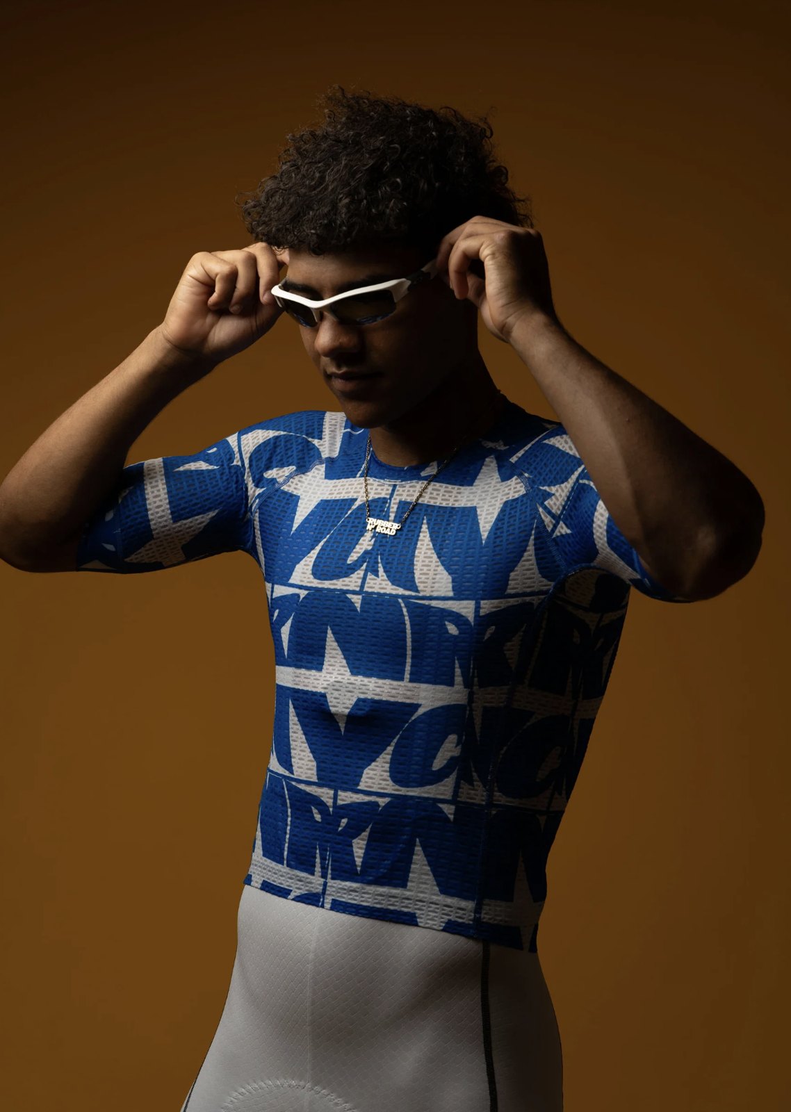 Enthusiasts persona reference: a young cyclist lifting his sunglasses, wearing a bold graphic-print short-sleeve jersey and white bib shorts